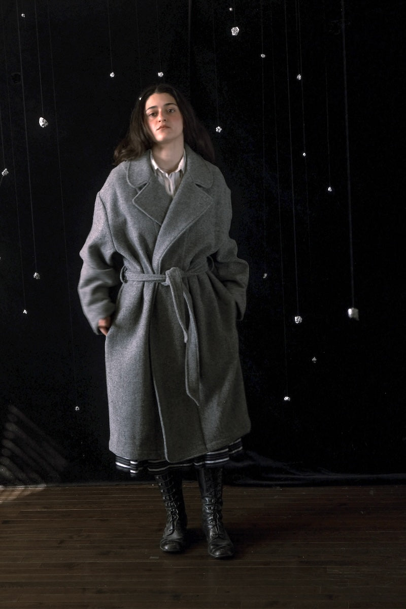 A woman stands in a studio wearing a long gray belted coat over a white collared shirt, with black lace-up boots. The background is black with hanging silver bead-like decorations and a wooden floor.