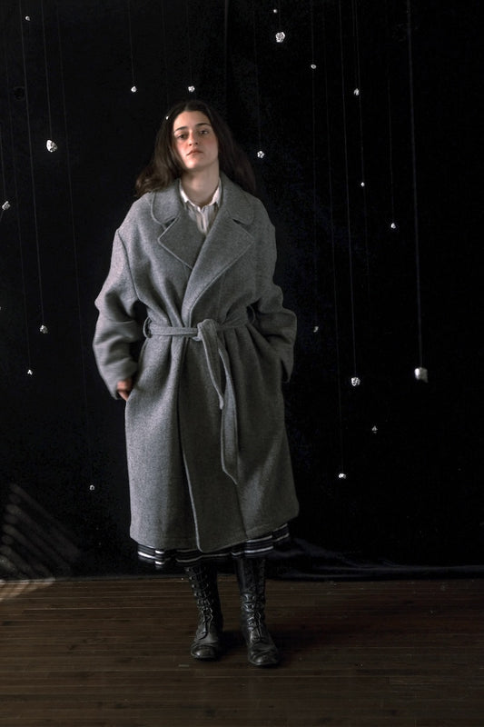 A woman stands in a studio wearing a long gray belted coat over a white collared shirt, with black lace-up boots. The background is black with hanging silver bead-like decorations and a wooden floor.