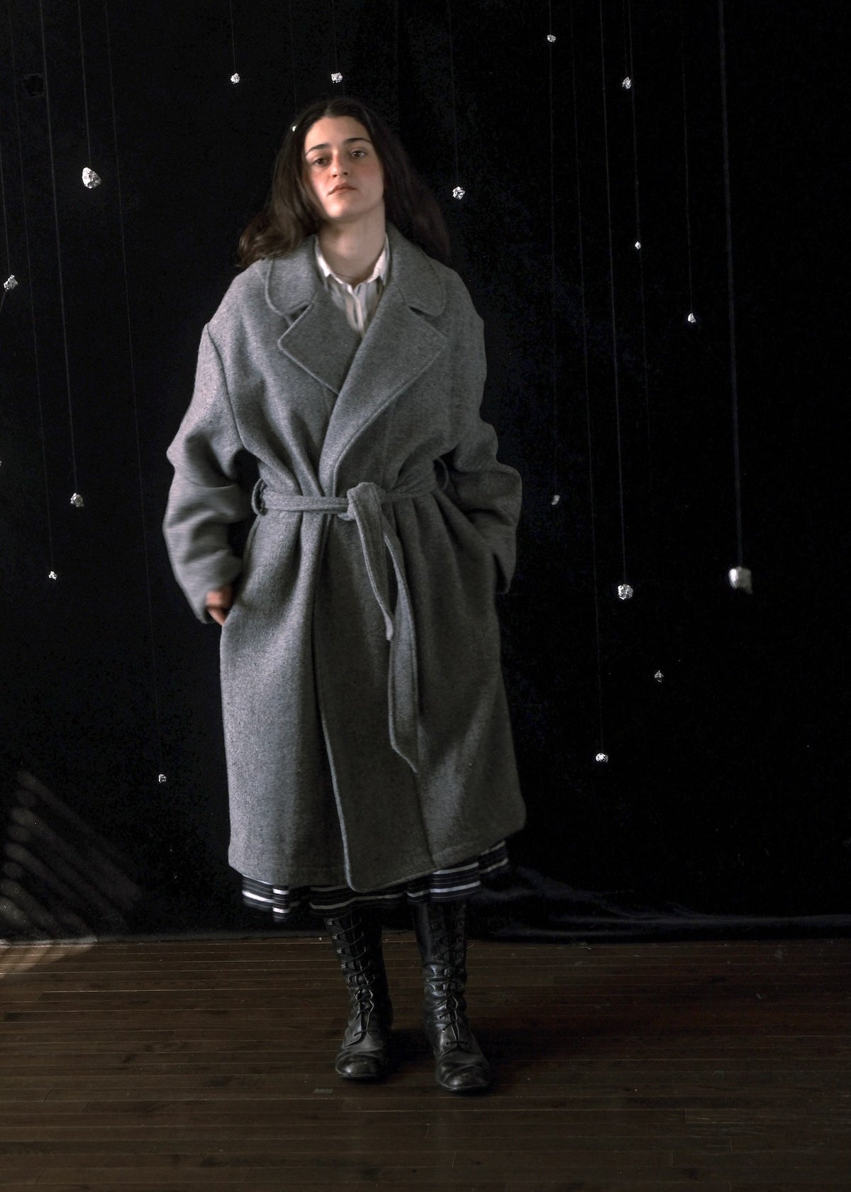 A woman stands in a studio wearing a long gray belted coat over a white collared shirt, with dark lace-up boots, against a black backdrop with hanging reflective lights.