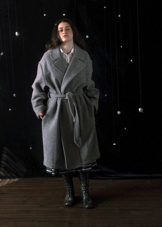 A woman stands in a studio wearing a long gray belted coat over a white collared shirt, with dark lace-up boots, against a black backdrop with hanging reflective lights.