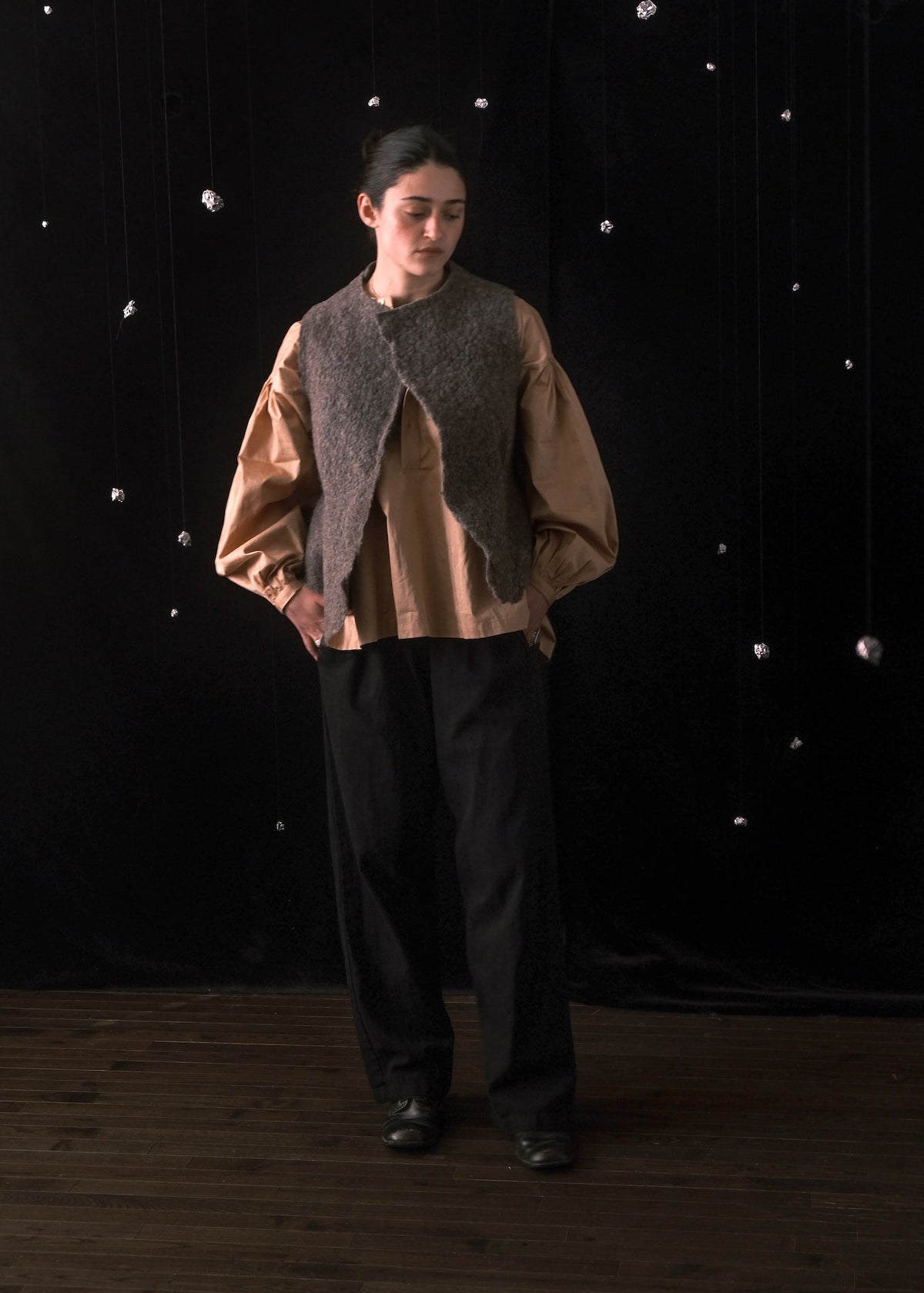 A model stands against a black fabric backdrop with small silver hanging ornaments, wearing a tan long-sleeve blouse with voluminous sleeves, a dark gray textured vest, and loose black trousers.