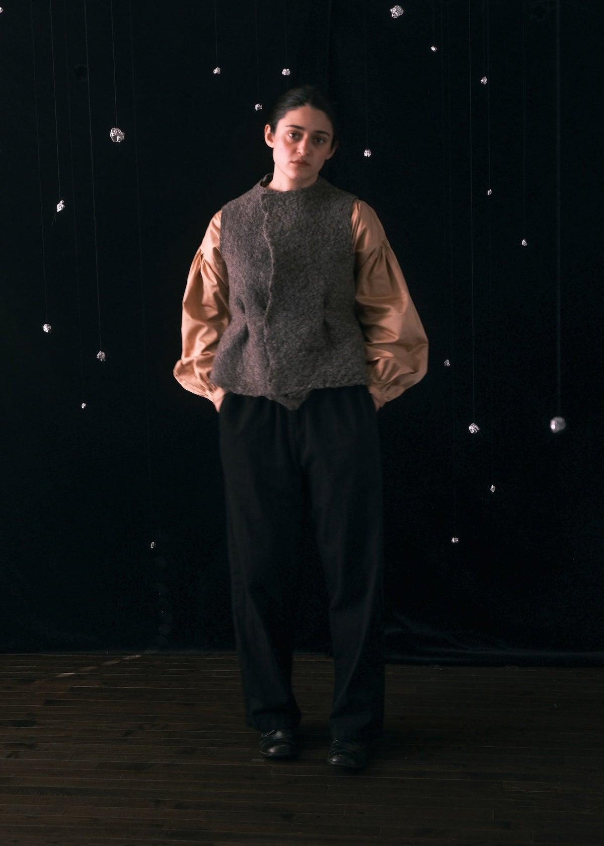 A model stands against a dark black fabric backdrop with hanging silver balloon-like decorations, wearing a brown textured knit vest over a light brown puff-sleeve blouse and black wide-leg trousers.