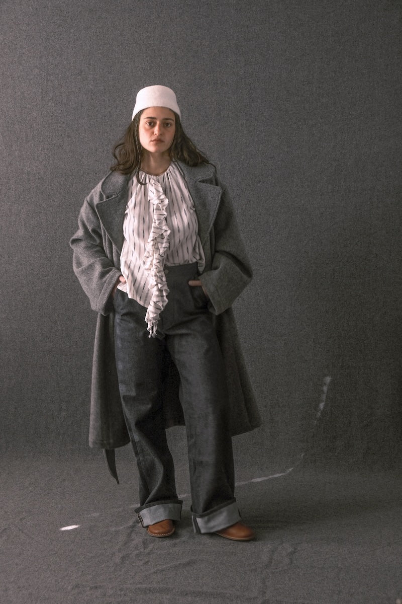 A model stands against a dark gray studio backdrop wearing a long charcoal gray coat over a white ruffled blouse, dark gray wide-leg trousers, a white cap, and brown shoes.