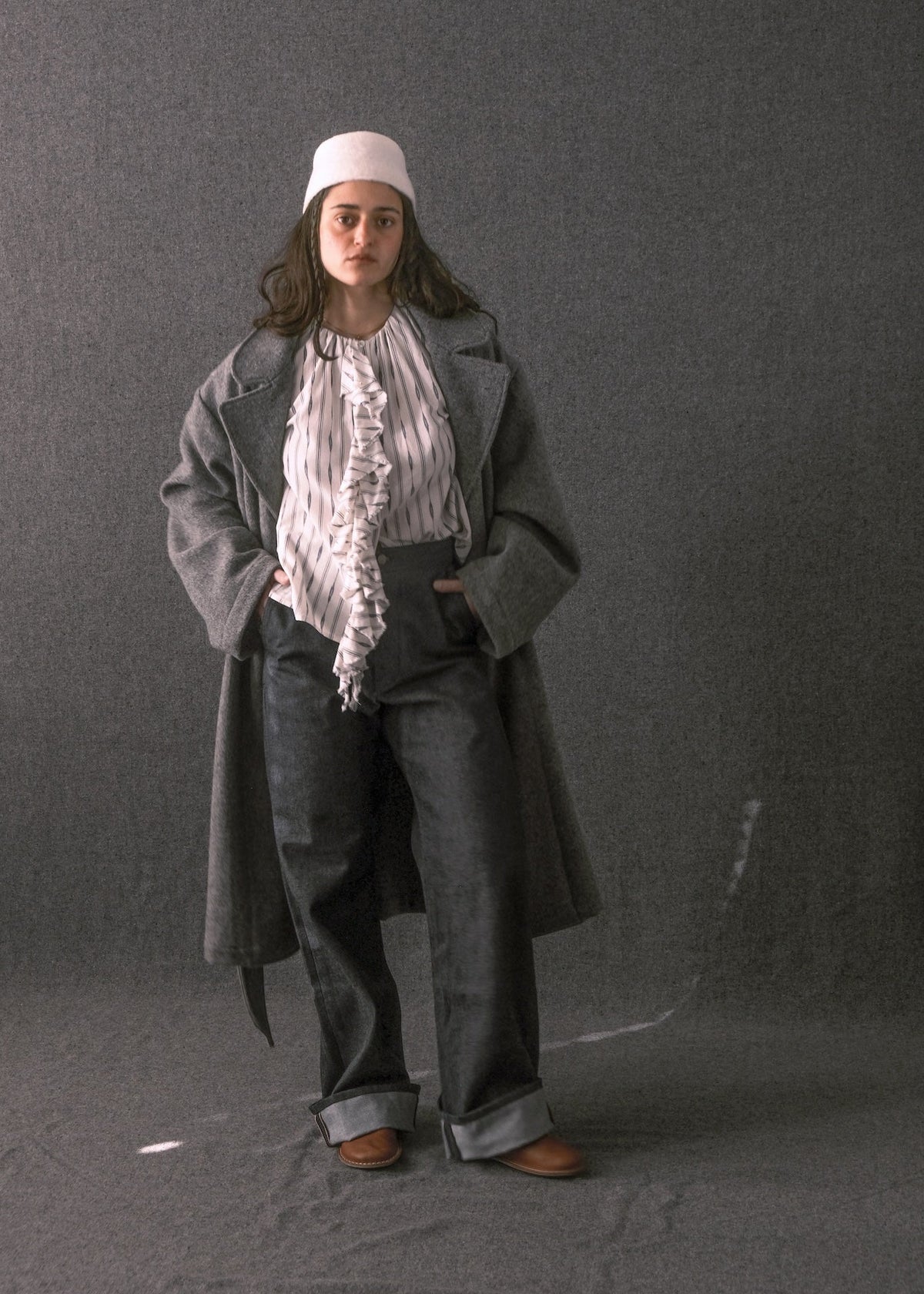A model stands against a dark gray studio backdrop wearing a long gray coat over a white ruffled blouse, dark wide-leg trousers, a white cap, and brown shoes.