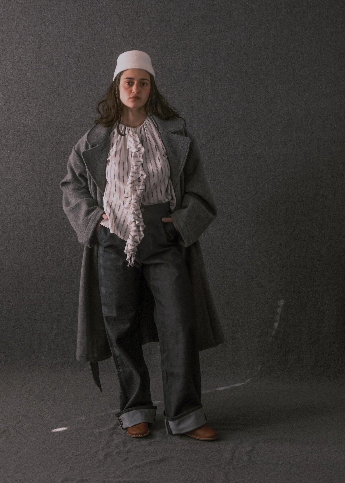 A model stands against a dark gray studio backdrop wearing a long charcoal gray coat over a white ruffled blouse, dark gray wide-leg trousers, a white cap, and brown shoes.
