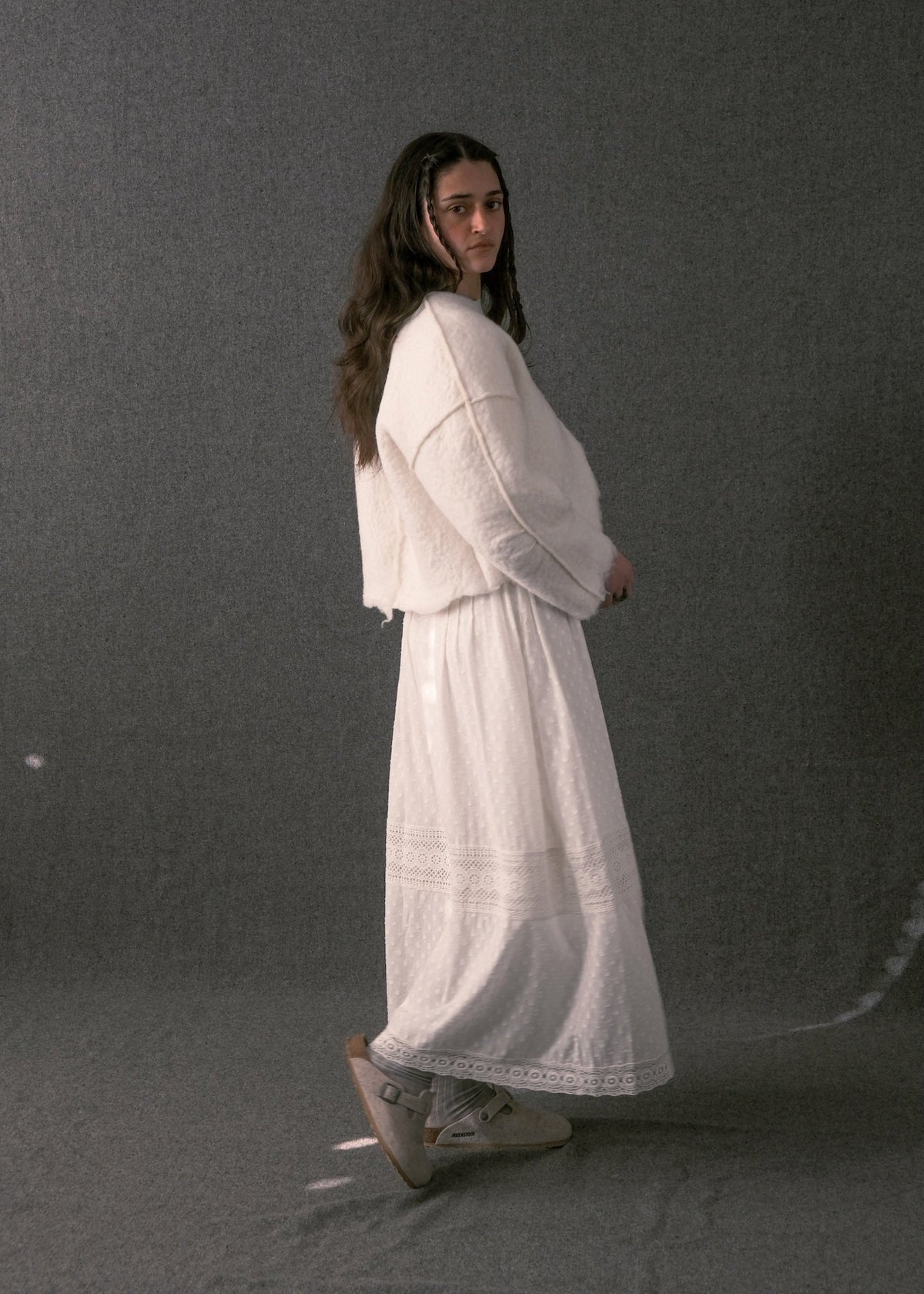 A woman wearing a white long-sleeve cropped cardigan and a matching long white skirt with lace trim, standing against a dark gray studio backdrop.