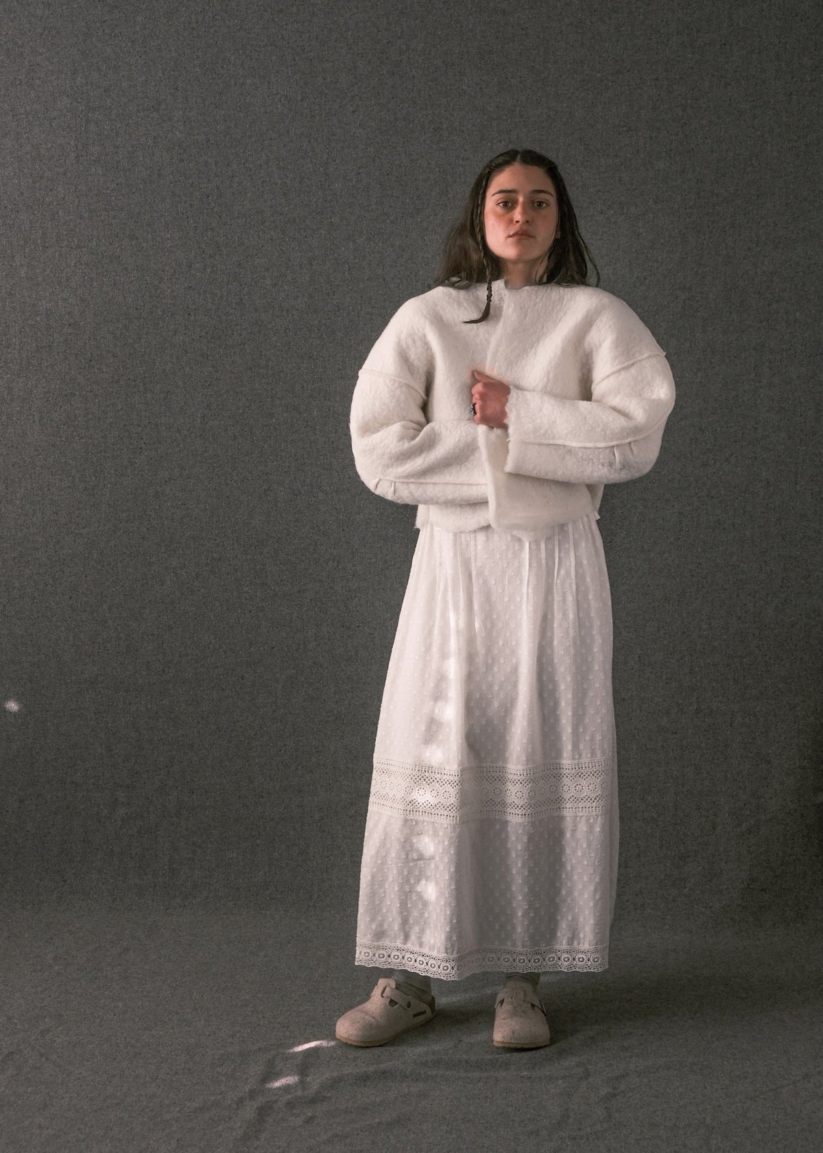A woman stands against a dark gray studio background wearing a voluminous white knit sweater and a long white skirt with lace trim, paired with beige shoes.