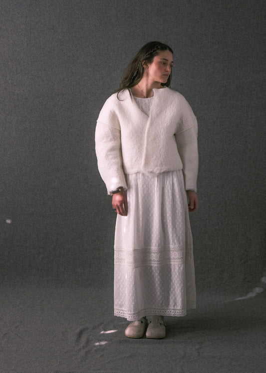 A model stands against a dark gray studio backdrop wearing a white fuzzy cropped jacket with long sleeves, a matching white textured maxi skirt, and light-colored slip-on shoes.