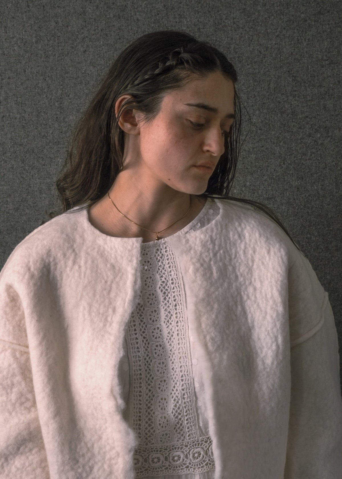 A woman wearing a cream-colored, textured knit cardigan over a white lace-trimmed top, set against a dark gray textured wall.