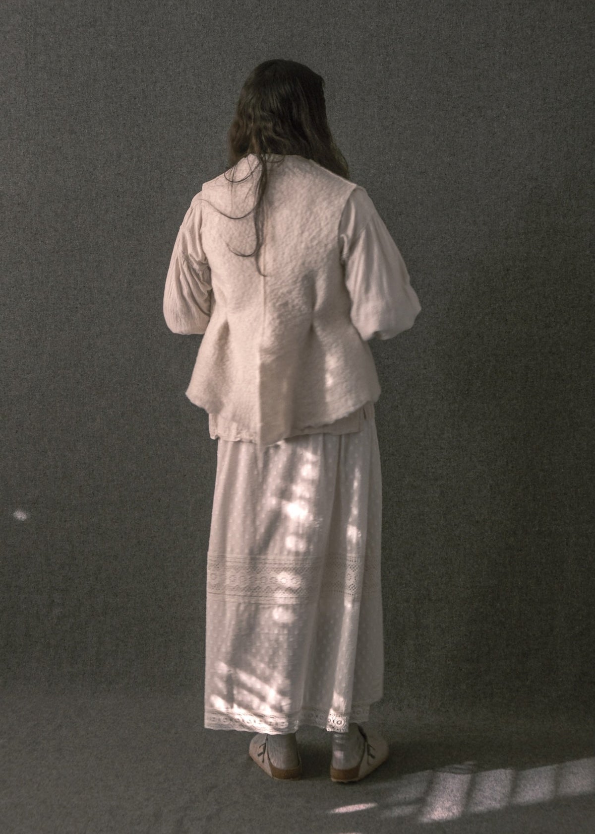 A woman stands with her back to the camera wearing a light beige textured long-sleeve top and a matching long skirt, set against a dark gray textured backdrop with dappled light on the floor.