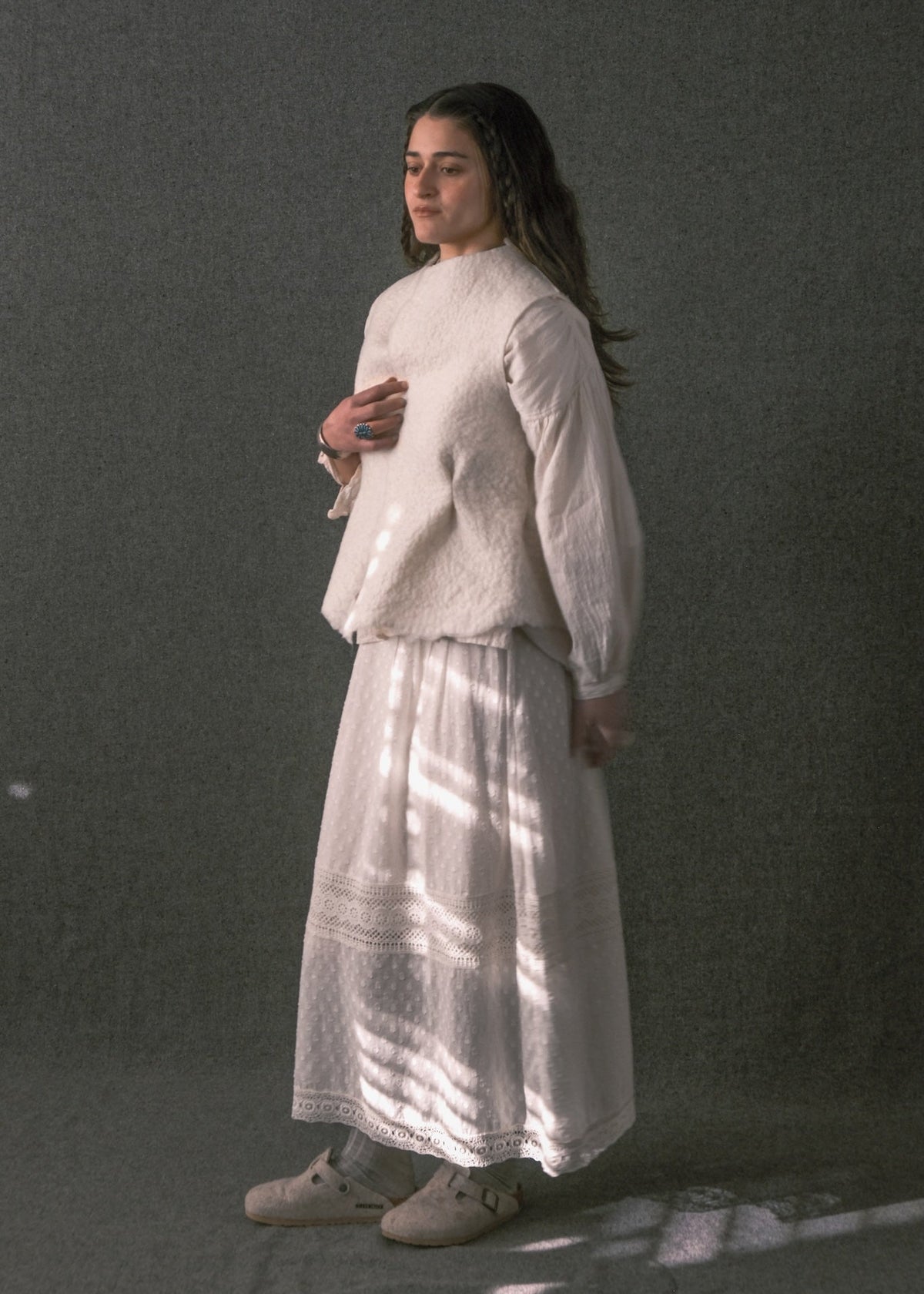 A woman stands in profile wearing a loose white long-sleeve top and a long white skirt against a dark gray studio backdrop, with dappled light casting shadows across her clothing.