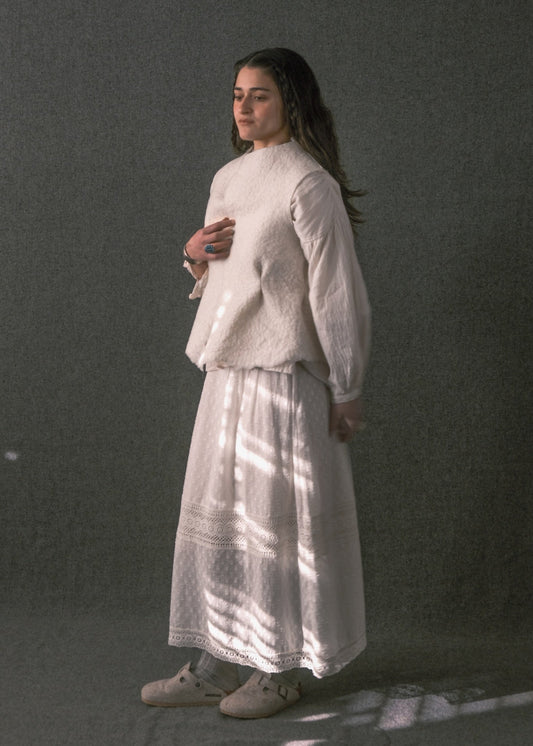 A woman stands in profile wearing a loose white long-sleeve top and a long white skirt against a dark gray studio backdrop, with dappled light casting shadows across her clothing.