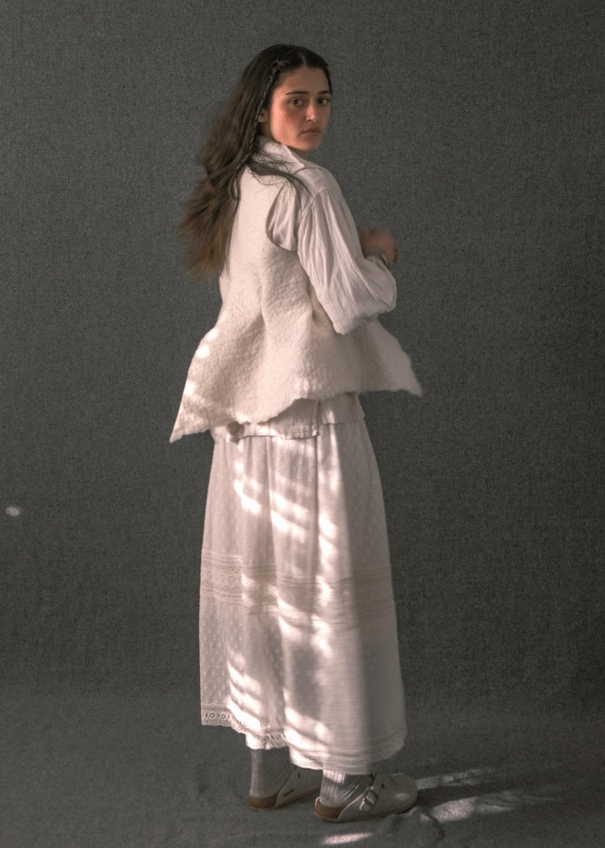 A woman stands in a studio wearing a white textured vest over a white long-sleeve blouse and a long white skirt, with gray socks and white sneakers. The background is a dark gray studio backdrop with dappled light patterns.