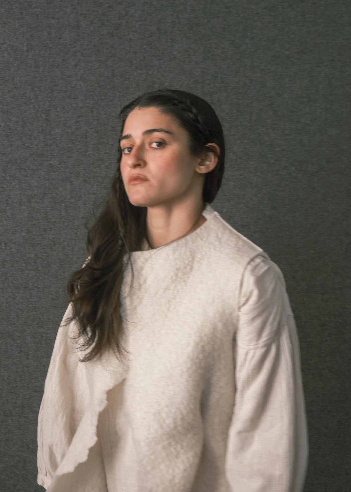 A woman with long dark hair wearing a textured off-white long-sleeve top, photographed against a dark gray textured studio background.