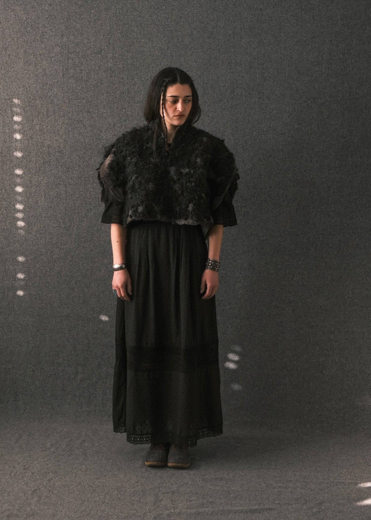 A model stands against a dark gray textured backdrop wearing a black textured cropped jacket with dramatic ruffled sleeves and a long black skirt.