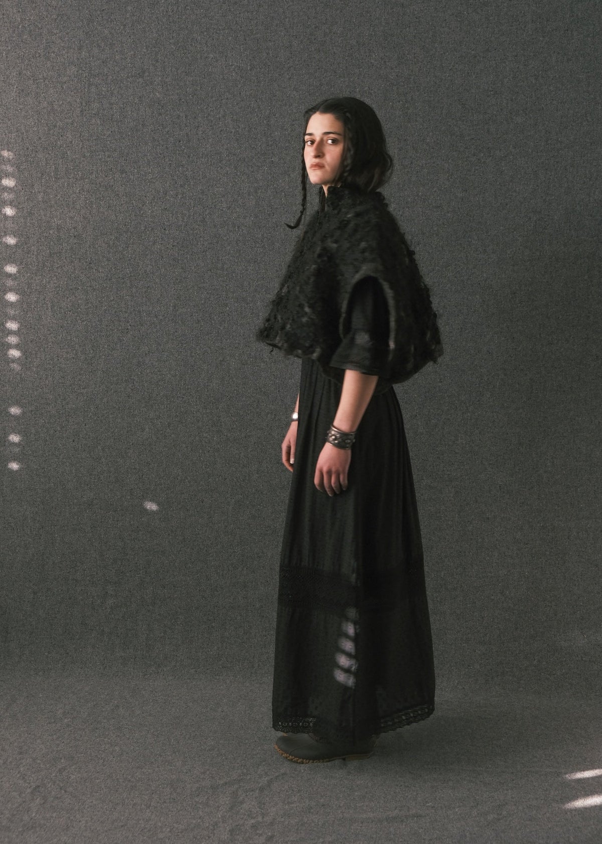 A model stands in profile wearing a long black dress with a textured black shawl or cape detail on the shoulders, posed against a dark gray studio backdrop.