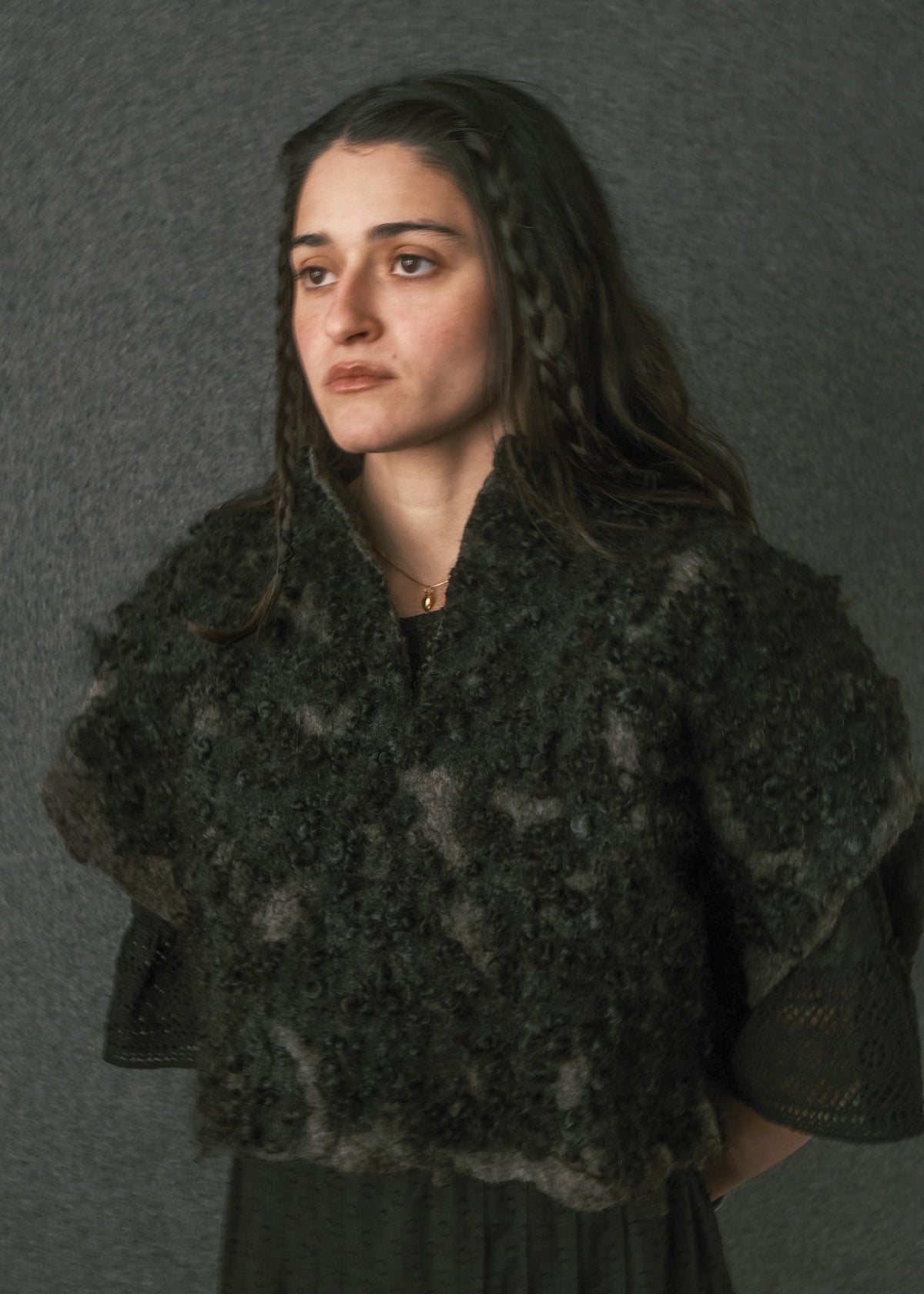 A woman wearing a dark, textured, shaggy knit or felt cape-style top with wide draped shoulders, photographed against a plain gray studio background.