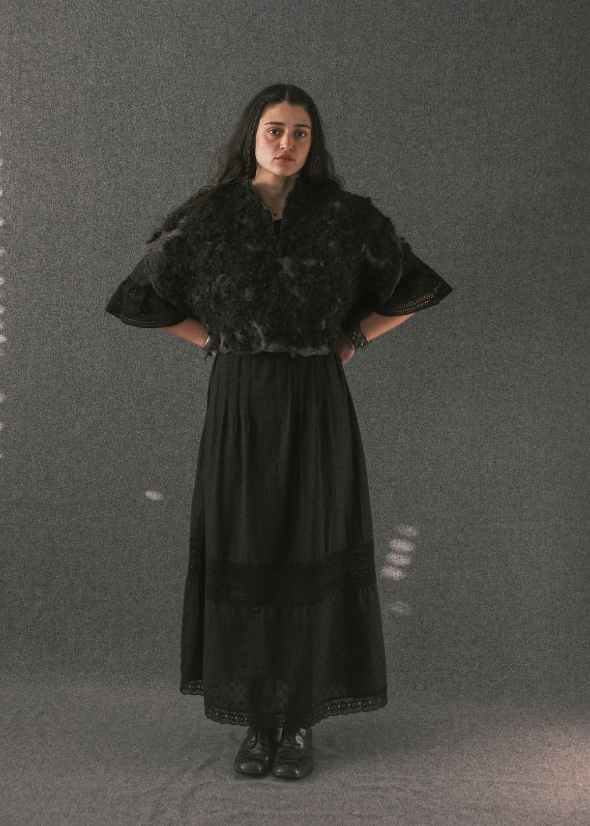 A woman stands against a textured gray studio backdrop wearing a long black dress with a dark textured shawl or capelet on the upper half, black lace trim at the waist and hem, and black shoes.