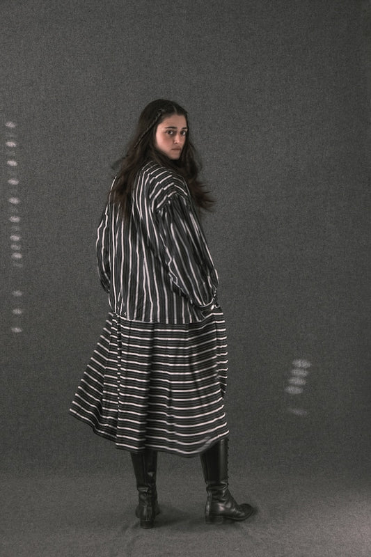 A woman wearing a black and white vertically striped long-sleeve coat with a matching striped skirt, paired with black leather boots, standing against a dark gray studio backdrop.