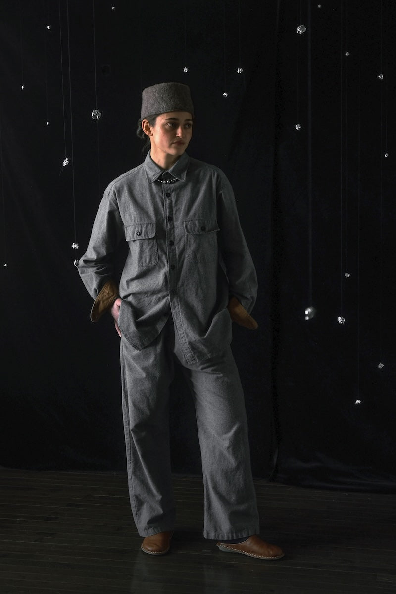 A person wearing a dark gray utility jumpsuit, matching gray cap, and brown boots stands against a black backdrop with hanging silver ornaments.