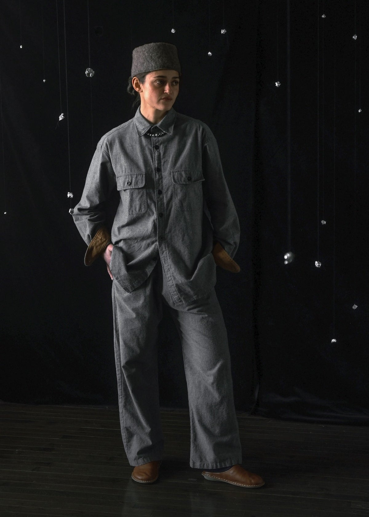 A person stands in a studio wearing a loose gray two-piece work uniform with a matching gray cap, brown gloves, and brown shoes against a black backdrop with hanging reflective dots.