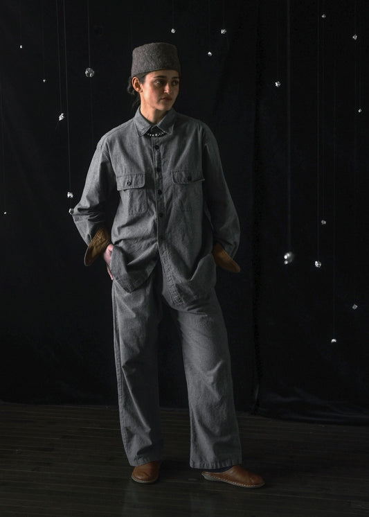 A person stands in a studio wearing a loose gray two-piece work uniform with a matching gray cap, brown gloves, and brown shoes against a black backdrop with hanging reflective dots.