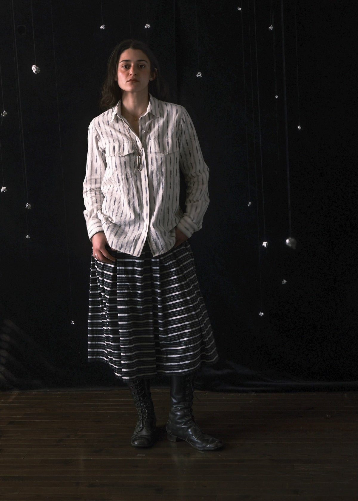 A model stands against a dark, starry backdrop wearing a white button-up shirt with thin vertical black stripes, a black and white horizontally striped midi skirt, and black leather boots.
