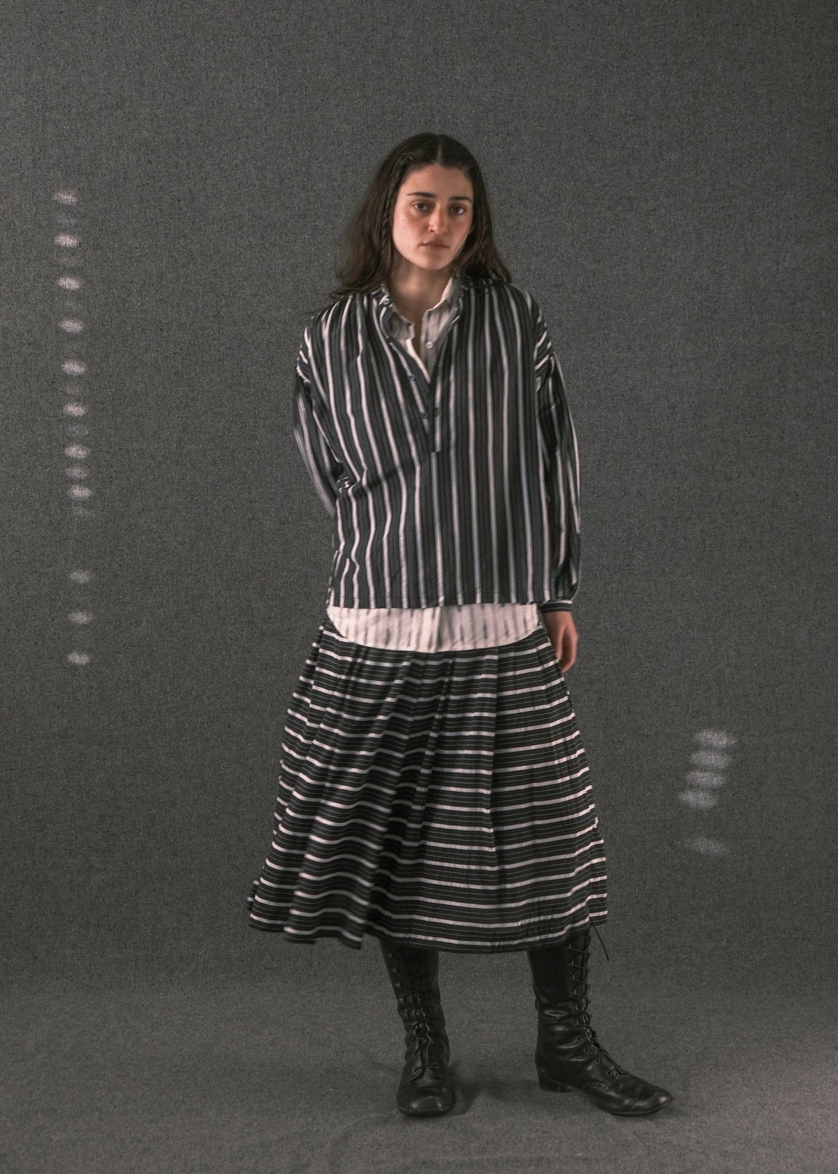 A woman stands against a dark gray studio backdrop wearing a black-and-white vertically striped long-sleeve shirt, a matching striped midi skirt, a light inner layer peeking out at the hem, and black lace-up boots.