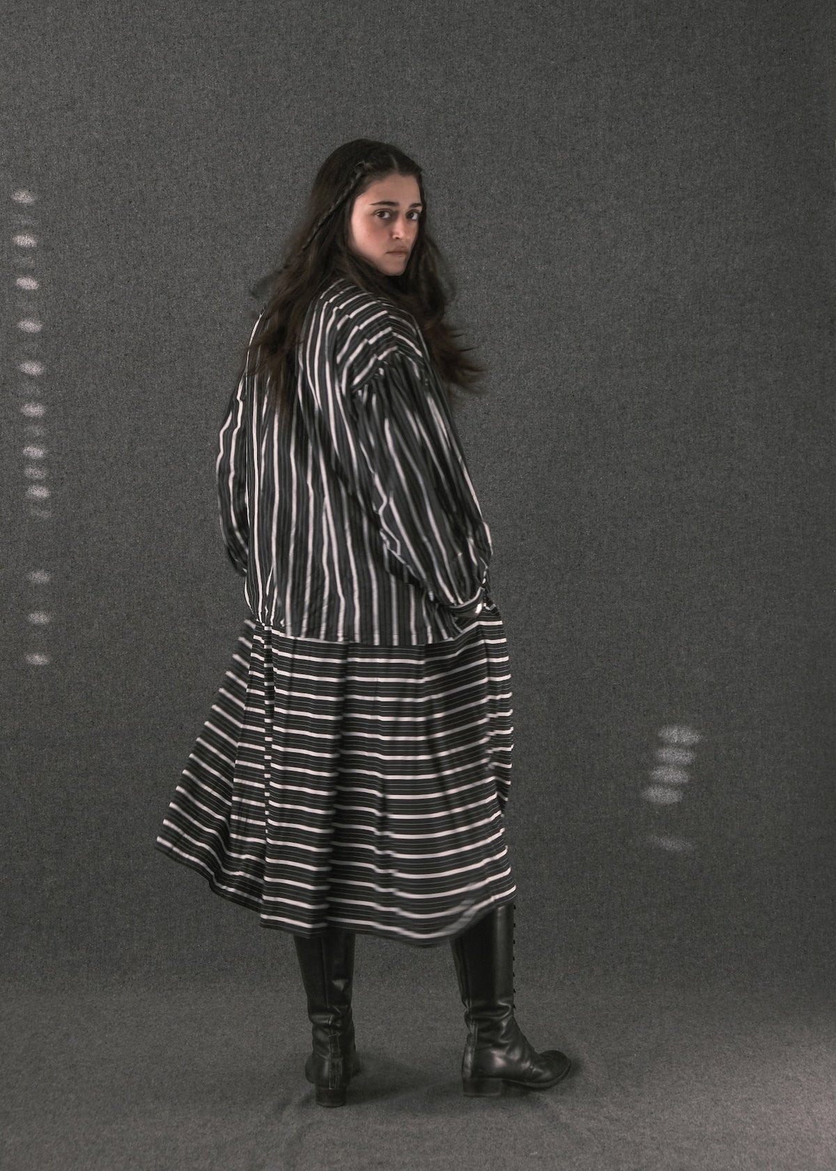A woman wearing a black and white vertically striped long-sleeve tunic dress with a flared skirt, paired with black leather boots, standing against a dark gray textured studio backdrop.