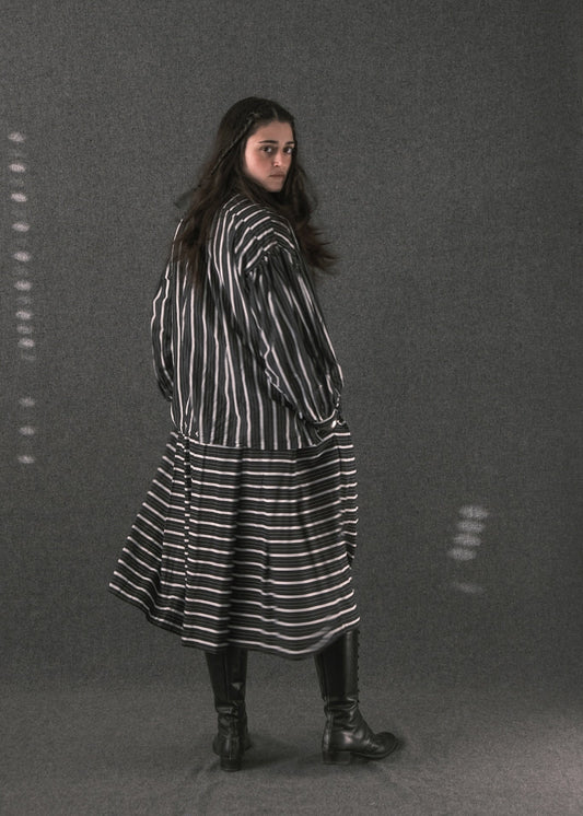 A woman wearing a black and white vertically striped long-sleeve tunic dress with a flared skirt, paired with black leather boots, standing against a dark gray textured studio backdrop.