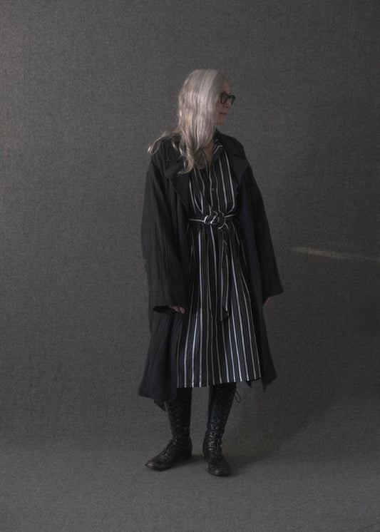 A person with long silver hair and sunglasses stands in a studio wearing a long black coat over a black-and-white vertically striped dress, with black lace-up boots, against a dark gray backdrop.