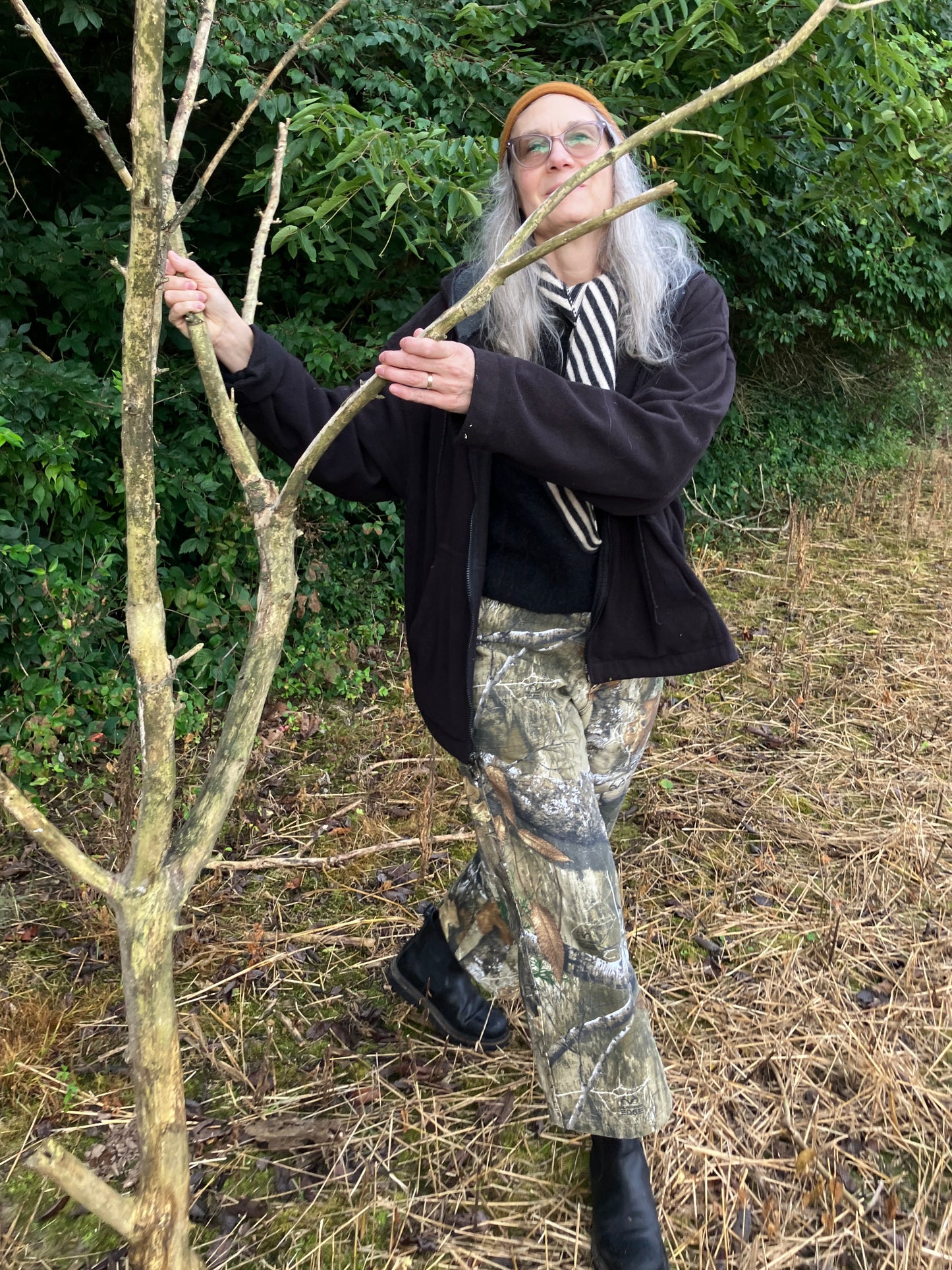 Person in outdoor setting holding a branch, wearing a black coat and camouflage pants.